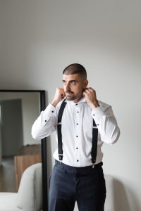 A man in a white dress shirt and black suspenders adjusts his collar while standing indoors, preparing for a wedding. A mirror and modern furniture are visible in the background, capturing the scene as if through the lens of a photographer.