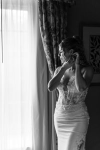 A bride in a lace wedding dress stands by a window, looking outside while putting on earrings. Natural light filters through sheer curtains, illuminating her face and gown—capturing the timeless elegance that every wedding photographer seeks in an editorial moment.