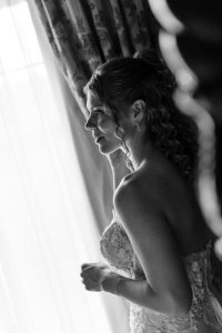 A woman in a wedding dress stands by a window, softly lit by natural light, looking outside. The black and white photography highlights her curly hair and lace gown, capturing an elegant moment that any Toronto photographer would cherish.