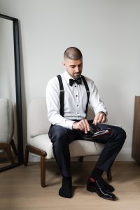 Man in formal attire with suspenders and bow tie sits on a chair, tying his dress shoe, as if preparing for a wedding. A mirror leans against the wall beside him, subtly reflecting the quiet anticipation of the moment. The calm, minimalist setting makes this scene ideal for an editorial photographer seeking refined simplicity.