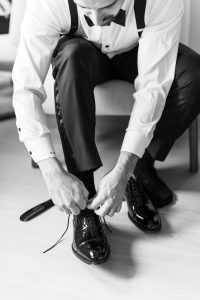 A man in formal attire sits on a chair and ties the laces on his shiny black dress shoes, perhaps preparing for a wedding. The image is black and white, with only his upper body and legs visible, giving it an editorial feel.