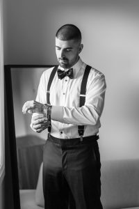 A man in formal attire with suspenders and a bow tie adjusts his wristwatch while standing beside a window, captured by a wedding photographer. A mirror and a sofa are visible in the background. The image is in black and white.