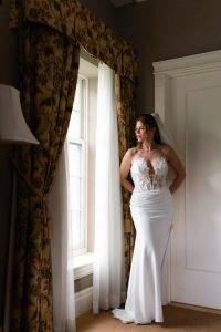 A bride in a white lace wedding dress stands by a window with floral curtains, looking outside. Soft sunlight illuminates her as she holds her hands behind her back, capturing a timeless moment that any documentary photographer would treasure.