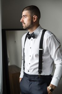 A man in formal attire with a bow tie and suspenders stands indoors, looking out a window with his hands in his pockets, as if waiting for the wedding photographer to capture the moment. A mirror and wooden furniture are in the background.