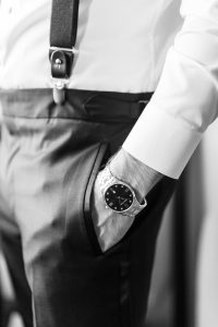 A person, possibly a wedding photographer, wearing dress pants, suspenders, a white shirt, and a wristwatch has their hand in their pocket. The image is black and white, focusing onto the hip and arm area.