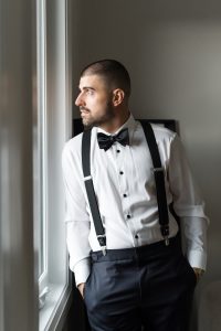 A man in a white dress shirt, black bow tie, and suspenders stands by a window, looking outside with his hands in his pockets, captured elegantly in this wedding photography scene.