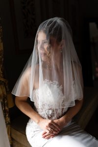 A bride in a lace wedding dress and veil sits on a bench, smiling and looking out a sunlit window, hands gently folded in her lap—the perfect moment of wedding photography captured by a talented photographer.