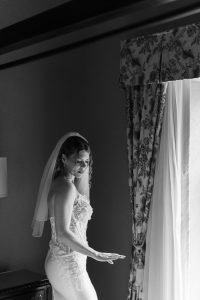 A bride in a strapless wedding gown and veil stands by a window with patterned curtains, softly lit by natural light, looking down with a peaceful expression. Captured in classic black and white, this editorial photograph embodies timeless elegance and the artistry of Toronto photography.