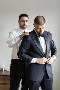 A man in a tuxedo adjusts his jacket while another man in a dress shirt stands behind him, resting his hands on his shoulders, helping him get ready—a classic moment of wedding photography captured with an editorial flair.