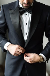 A man in a black tuxedo adjusts his jacket button, captured through skilled photography. He wears a white shirt, black bow tie, and a gold wristwatch, with his head slightly bowed and face partially visible.