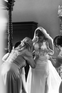 A bride in a lace wedding gown and veil stands as two women adjust her dress, preparing her for the ceremony in a softly lit room, while a photographer from Toronto captures the intimate moment.