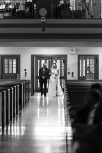 An editorial photograph captures a bride in a wedding gown walking down the aisle with an older man in a suit, likely her father, in a church with empty pews. The scene is rendered beautifully in black and white.