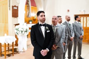 A groom in a black tuxedo stands at the altar with his groomsmen in grey suits behind him, waiting inside a church decorated with white flowers—a timeless moment beautifully captured by a Toronto photographer in an editorial style.