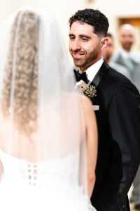 A groom in a black tuxedo smiles at his bride, who is wearing a white dress with a veil, during a wedding ceremony, beautifully captured in documentary photography style in Toronto.