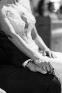 A bride and groom sit side by side, holding hands. The bride wears a lace dress and an engagement ring is visible on her finger. Captured in black and white by a Toronto photographer, the photo has a timeless, documentary feel.