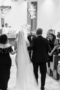 A bride in a long veil walks down the aisle with two people, approaching the groom at the altar in a church. Guests look on, smiling and taking photos as this heartfelt wedding moment unfolds. Captured in black and white, the scene has an elegant editorial quality.