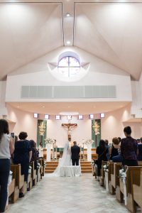 A bride and groom stand at the altar in a church, facing a priest for their wedding ceremony. Guests are seated in pews, watching as the photographer captures every heartfelt moment. The church has high ceilings and a large crucifix at the front.
