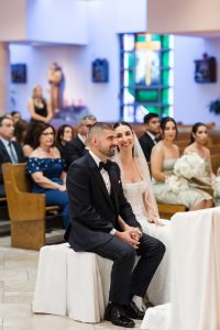 A bride and groom sit together at the altar in a church, smiling during their wedding ceremony as the photographer captures the special moment. Guests are seated in pews behind them, and stained glass windows and statues are visible in the background.