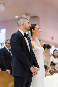 A bride and groom stand side by side, holding hands during a wedding ceremony as a photographer captures the moment. The groom wears a black tuxedo and the bride wears a white lace dress with a veil. People are visible in the softly blurred background.