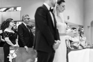 Black and white editorial photography capturing wedding guests sitting and standing in pews, facing forward. A bride and groom stand in the foreground, out of focus, while several people look on attentively in the background.