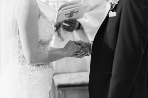 A bride and groom hold hands during a wedding ceremony, with an officiant reading from a book in the background. Captured in black and white, this image reflects the genuine emotions often sought by a documentary wedding photographer.