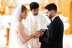 A bride and groom exchange rings during a wedding ceremony, with a priest standing behind them holding an open book, beautifully captured by a talented Toronto wedding photographer.
