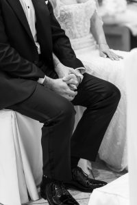 A couple sits side by side, holding hands. Captured with an editorial eye by the photographer, the man wears a dark suit and dress shoes; the woman wears a white, beaded gown. The black and white image is focused onto their clasped hands and seated posture.