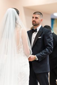 A groom in a black tuxedo holds hands with a bride in a white dress and veil during their wedding ceremony, both looking at each other, capturing a timeless moment of love through elegant editorial photography.