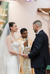A bride and groom hold hands and smile at each other during a wedding ceremony, with a priest standing between them, holding a microphone and a book, as the wedding photographer captures this editorial moment.