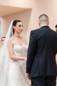A bride in a white wedding dress and veil holds hands with a groom in a dark suit during an editorial wedding ceremony, both standing and facing each other.