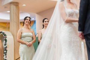 Two bridesmaids in light green dresses stand smiling and watching as a veiled bride, mostly out of focus, participates in an editorial-style wedding ceremony.