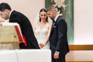 A bride and groom, both in formal attire, smile and look at the bride’s hand during a wedding ceremony inside a beautifully decorated venue. The moment is elegantly captured through wedding photography, as a person in a suit stands nearby holding an open book.