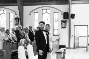A bride and groom stand at the front of a church, smiling, while guests seated in pews watch them. Captured in timeless black and white photography, this scene beautifully reflects the skill of a Toronto photographer.