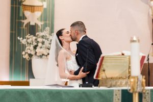 A bride and groom share a kiss at the altar during their wedding ceremony, beautifully captured onto film in this stunning display of wedding photography, with flowers and a large Bible visible in the church background.