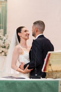 A bride and groom smile at each other and embrace at the altar during their wedding ceremony, with flowers and a book on a stand in the background, capturing an editorial-inspired moment of love and joy.