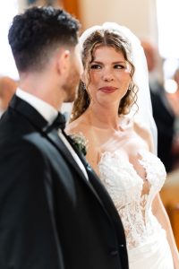 A bride in a white lace dress and veil smiles at the groom in a black tuxedo during their wedding ceremony in a bright indoor setting, beautifully captured by a talented Toronto photographer.