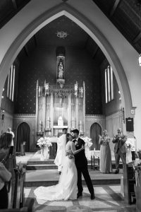 A bride and groom share a kiss at the altar in a church while wedding guests and the bridal party look on and applaud. Captured in classic black and white, this Toronto wedding moment has a timeless, documentary feel.