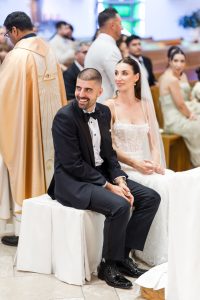 A bride and groom sit side by side during a wedding ceremony in a church, captured beautifully through wedding photography. The groom is smiling and looking away, while the bride looks forward. Guests and a priest in gold robes are in the background, adding onto the sense of joyous celebration.
