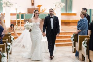 A bride and groom walk down the aisle, smiling and holding hands, as a photographer captures this joyful moment. The bride holds a bouquet of white flowers. Guests watch from pews. The setting is a decorated church altar—perfect for wedding photography.