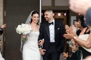 A bride and groom smile as they walk arm in arm, surrounded by applauding guests, while a photographer captures the joyful moment. The bride holds a bouquet of white flowers and wears a white dress and veil; the groom wears a black tuxedo.