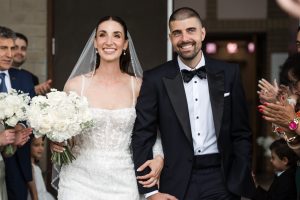 A bride in a white dress and a groom in a black tuxedo smile and walk arm in arm, holding a bouquet of white flowers, as people around them clap and celebrate—all beautifully captured by wedding photography.