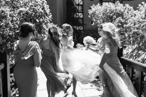 A bride in a white gown is surrounded by four bridesmaids in matching dresses, helping with her dress outdoors on a sunny day. The lively scene, full of smiles and conversation, perfectly captures the essence of wedding photography in Toronto.