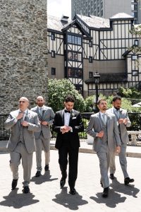 Five men in suits, one in black and four in gray, walk confidently outdoors near a stone building and Tudor-style architecture on a sunny day, as if captured by a documentary photographer at a stylish wedding.