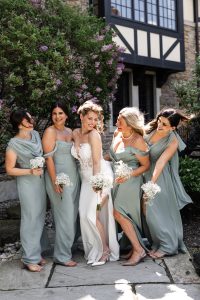 A bride in a white dress smiles with four bridesmaids in matching sage green dresses, holding small white bouquets, posing together outdoors near a flowering bush and a stone building, perfectly captured by an editorial photographer with a documentary style.