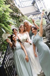 Five women in pastel dresses, including a bride in white, smile and hold small bouquets outdoors beside greenery and stone, celebrating together on a wedding day in a style reminiscent of editorial wedding photography.