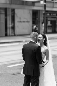 A bride and groom, dressed in wedding attire, stand facing each other on a city street in an editorial photography style. The bride holds a bouquet and wears a veil, while the blurred buildings fade into the background, drawing attention onto the couple. The image is black and white.