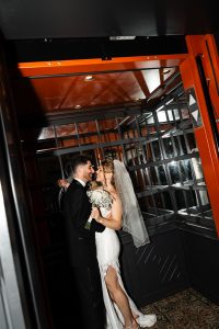A bride and groom stand closely together in an elevator, smiling at each other in a moment that feels both editorial and intimate. The bride wears a white dress and veil, holding a bouquet, while the groom is in a black tuxedo. Captured by the photographer in a modern setting with reflective walls, the scene has a refined, documentary quality.