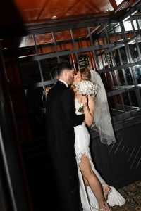 A bride in a white dress and veil hugs a groom in a black suit, holding a bouquet of white flowers, both smiling inside a modern, glass-paneled venue—captured with an editorial, documentary photography style.
