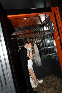 A bride and groom embrace and kiss in an elevator, captured beautifully by a talented Toronto photographer. The bride holds a bouquet and wears a veil and white dress, while the groom is in a black suit. The scene is warmly lit and intimate, highlighting the artistry of wedding photography.