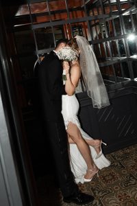 A bride in a white dress and veil holds a bouquet to her face while embracing a groom in a dark suit. They stand closely together indoors near a mirrored wall, beautifully captured by a Toronto wedding photographer.